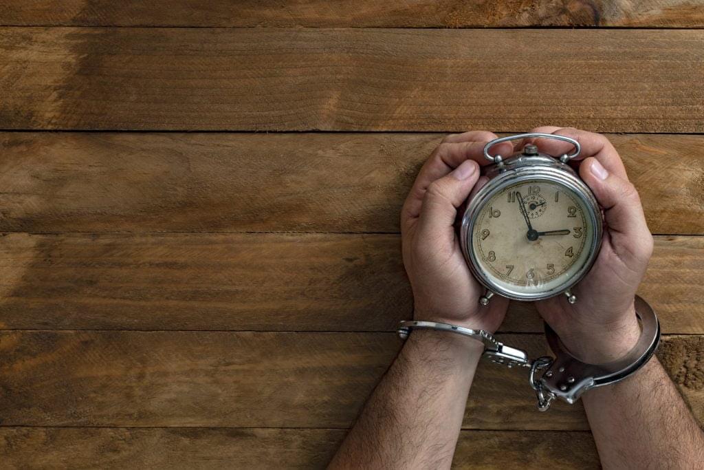 Time in jail. Shackled hands holding an alarm clock on a wooden table.
